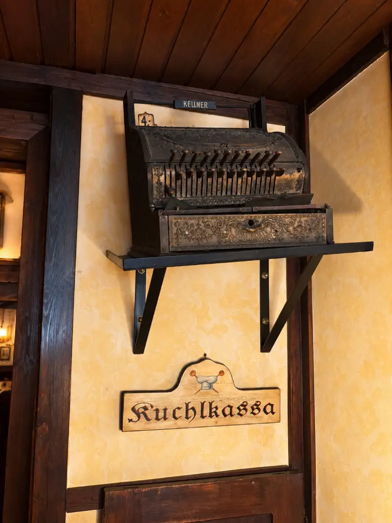 Alte Registrierkasse auf einer Wandkonsole, darunter das geschnitzte Schild „Kuchlkassa“